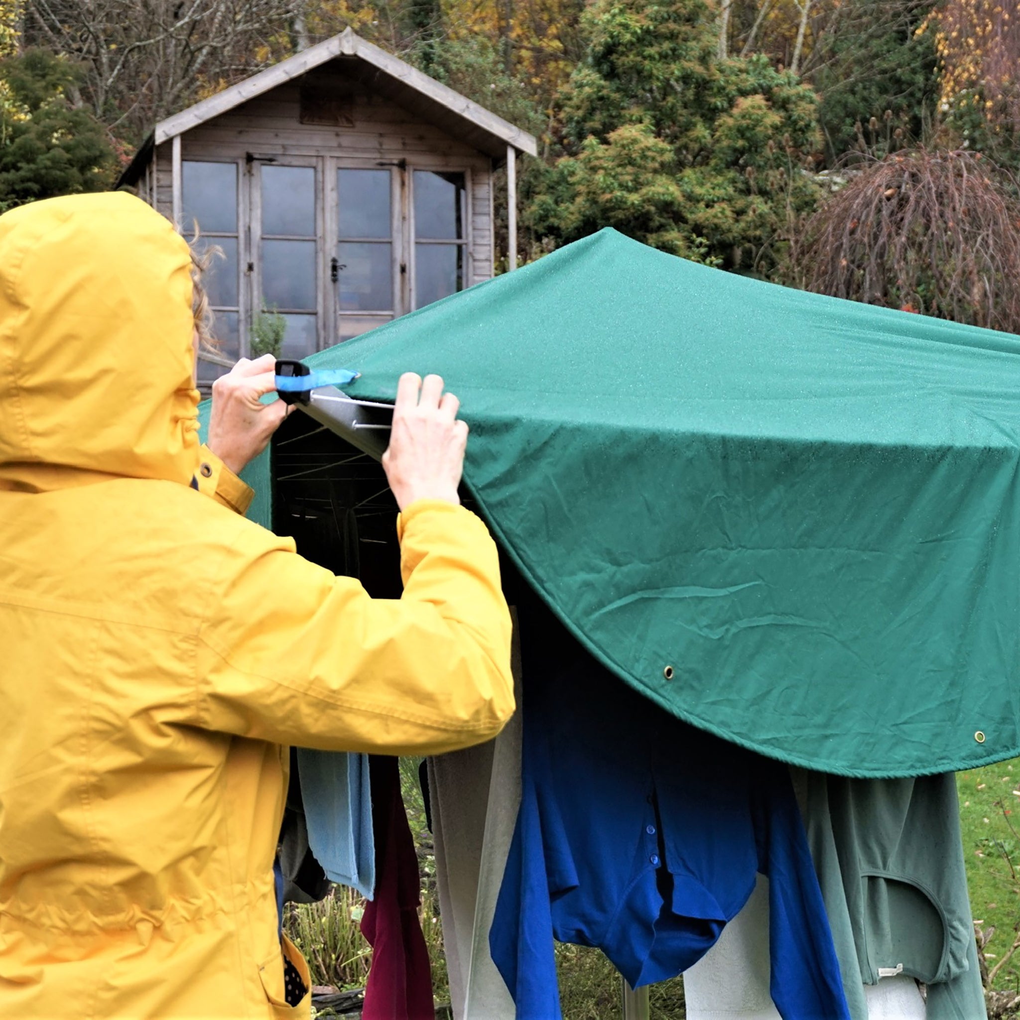 A woman wearing a yellow raincoat hangs laundry on a rotary dryer then covers it with a green Laundry Mac which protects the laundry from the rain