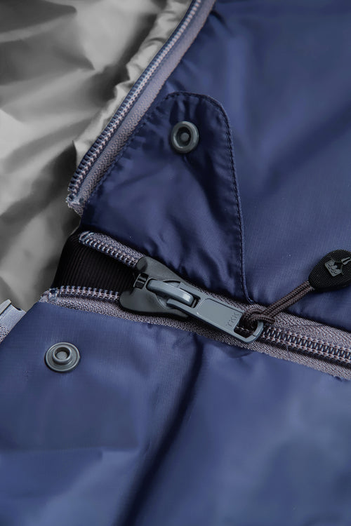A dark navy blue sleeping bag with a gray interior lining, shown vertically against a plain white background. The sleeping bag features a zippered pocket on the upper side and a small red and white logo near the bottom.