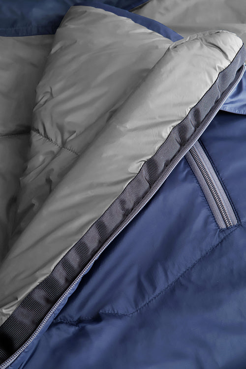 A dark navy blue sleeping bag with a gray interior lining, shown vertically against a plain white background. The sleeping bag features a zippered pocket on the upper side and a small red and white logo near the bottom.