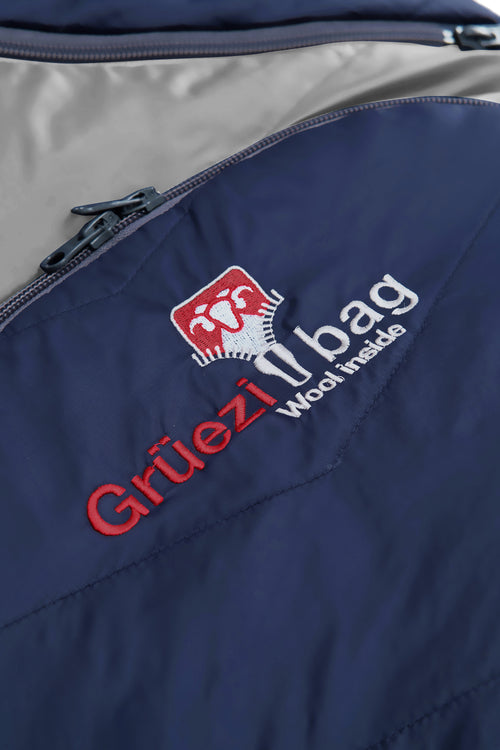 A dark navy blue sleeping bag with a gray interior lining, shown vertically against a plain white background. The sleeping bag features a zippered pocket on the upper side and a small red and white logo near the bottom.