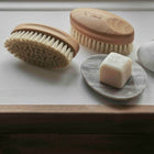 Two wooden brushes, a bar of soap on a stone, and a pink washcloth on a white surface.