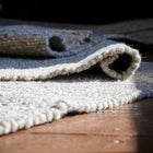 A close-up of a thick, textured wool rug in a light grey colour, shown partially rolled up on a rustic wooden floor.
