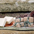 Three creamy white bars of olive oil castile soap, with three wrapped packages of soap, and a fircone in the background