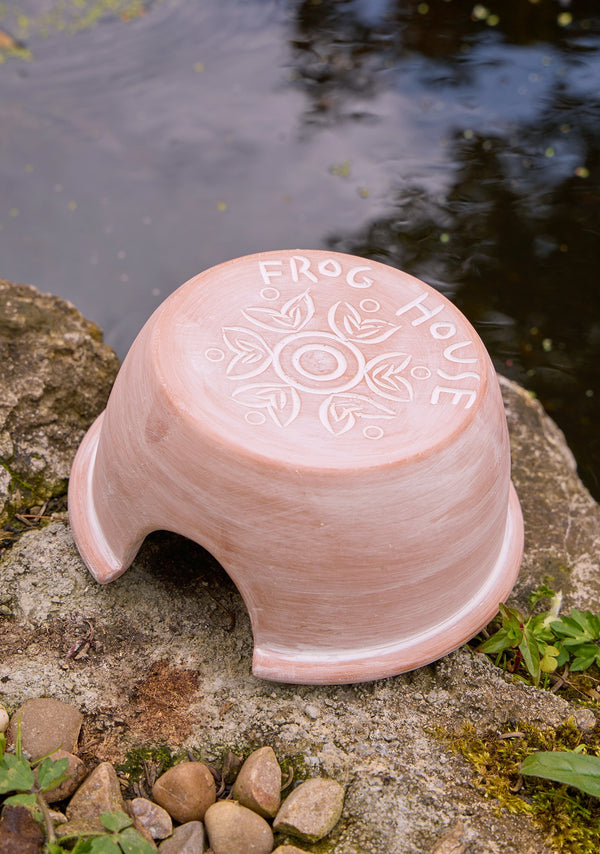 Ceramic frog house with 'Frog House' engraving on a stone surface near water.