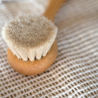 A close-up of a small wooden baby hair brush with soft beige bristles, resting on a woven beige fabric surface.