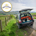 Car parked with lake district mountains in the background, with two dogs on roll-up dog bed unrolled in the back of the car, featuring 'Gift of the Year' award badge.