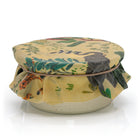 A  small pot covered with a beeswax fabric lid featuring a botanical pattern in green, blue, and orange on a yellow background, tied with a thin elastic band.