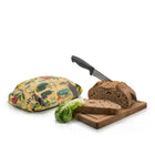 A reusable large beeswax wrap with a colourful jungle print sits next to a loaf of dark rye bread on a wooden cutting board with a knife and a green lettuce leaf, all on a white background.