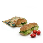 Two subway sandwiches with lettuce, one wrapped in a reusable patterned beeswax food bag and one unwrapped, with three red cherry tomatoes on a white background.