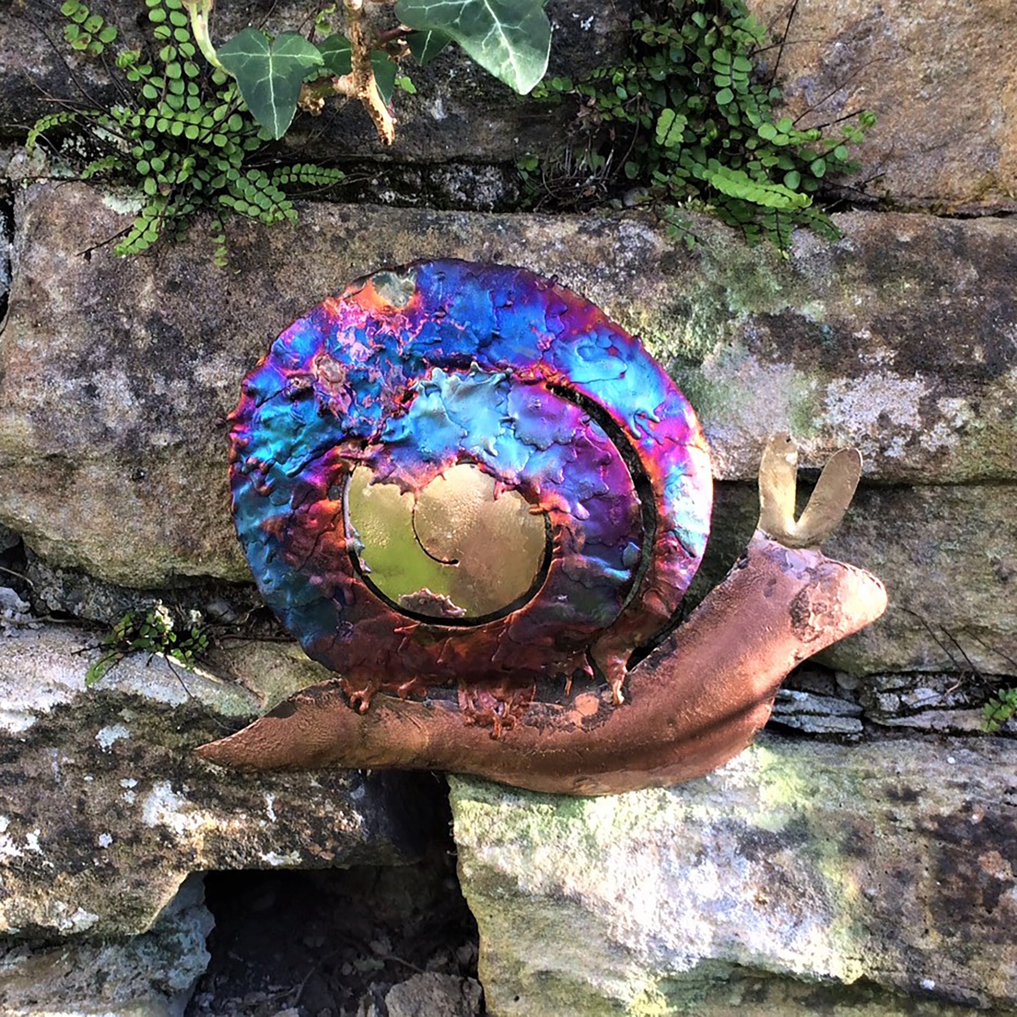 Copper ornamental snail with copper coloured body and iridescent blues, purples, on the curly shell, placed on the garden wall.