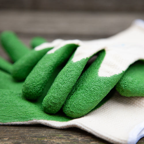 Gardening gloves with green fingertips on a wooden surface with gardening tools in the background.