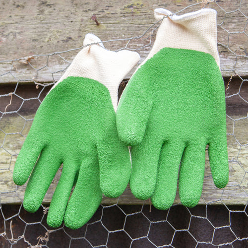 Gardening gloves with green fingertips on a wooden surface with gardening tools in the background.